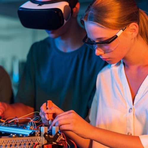 Group of teens doing experiments in robotics in a laboratory. Boy and girls in protective and VR glasses working with a robot. Red and blue illumination